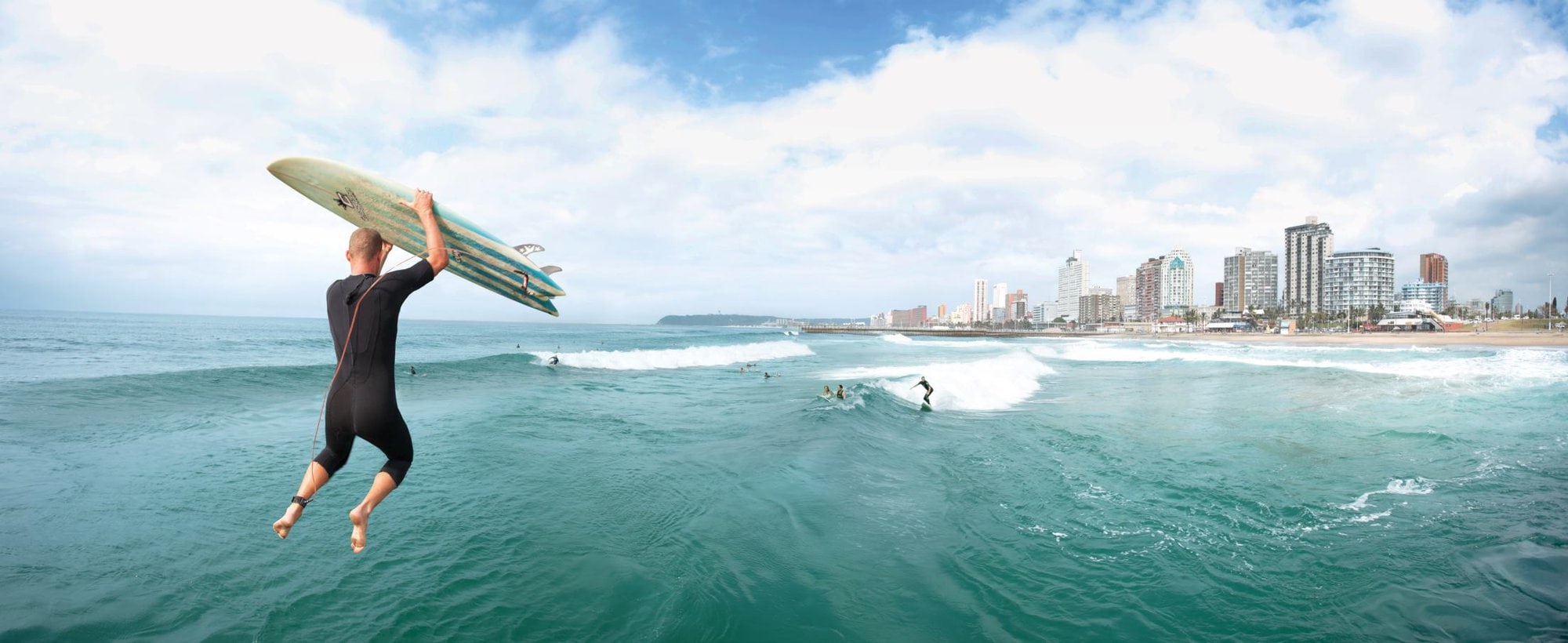 Een surfer die van de pier in het water springt met op de achtergrond de skyline van Durban