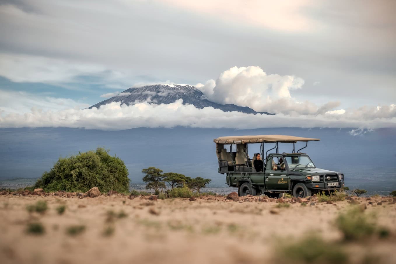 Besneeuwde top van Kilimanjaro steekt uit boven nevelig wolkendek met op de voorgrond bezoekers in een safari-jeep in Amboseli